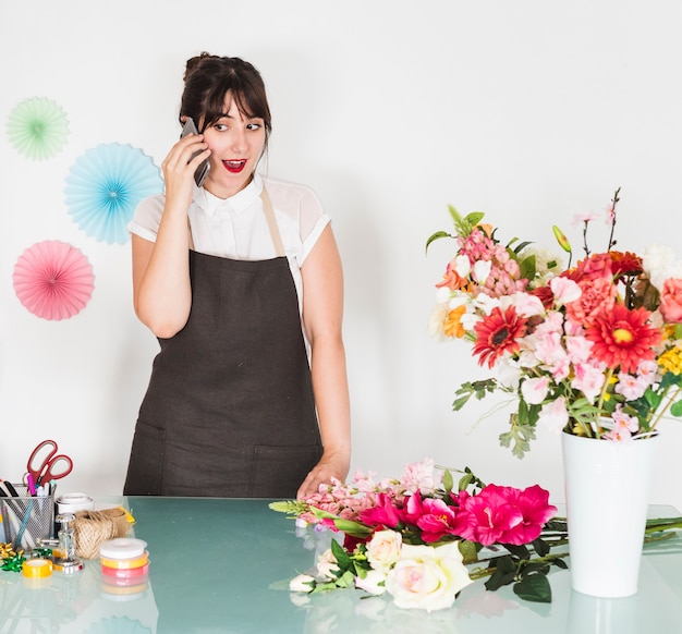 Überraschte Frau, die auf Smartphone mit Blumenstrauß auf Schreibtisch spricht