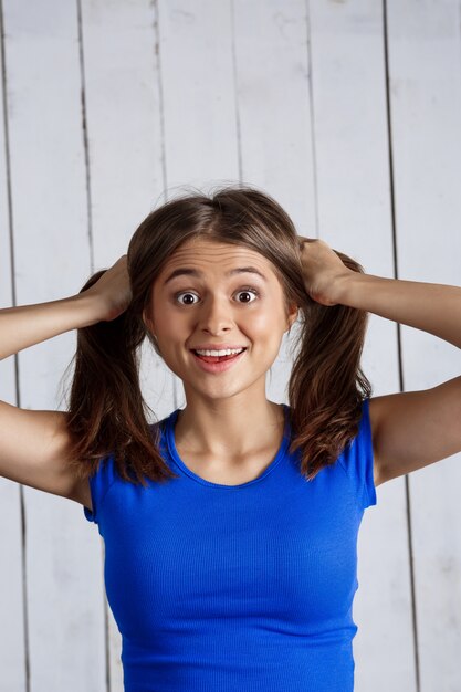 Überraschte Frau, die Haare in den Händen über weißer Holzwand hält.