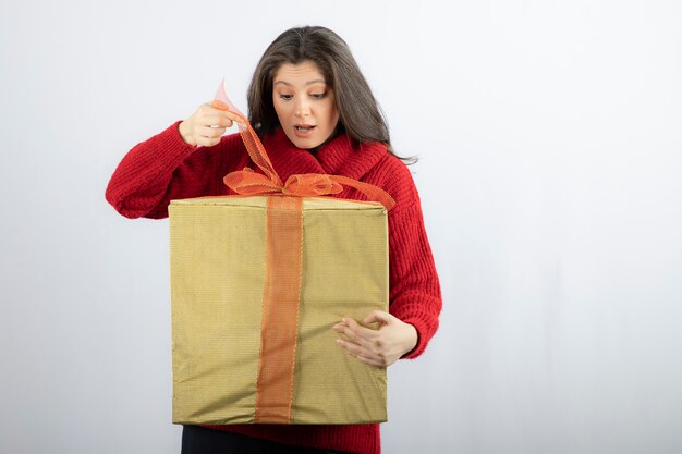 Überraschte Frau im roten Pullover, die eine Schachtel mit Weihnachtsgeschenk öffnet.