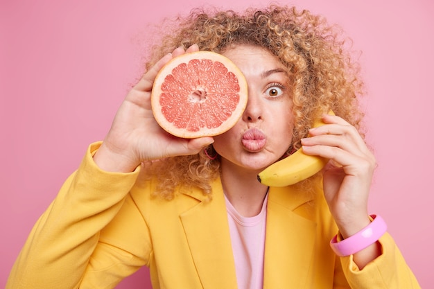 Überraschte Frau mit gefalteten Lippen, lockiges Haar hält die Hälfte der Grapefruit über dem Auge frische Banane in der Nähe des Ohrs, das Telefongespräch gibt vor, lockiges Haar in modischer gelber Jacke zu tragen. Weibchen hält reife Früchte