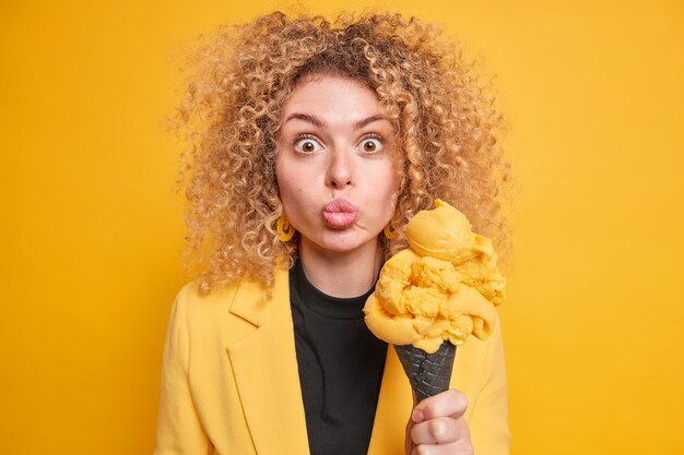 Überraschte Frau mit lockigem Haar hält die Lippen abgerundet hält appetitliches Eis isst kalt gefrorenes Dessert bei heißem Wetter trägt stilvolle Jacke isoliert über gelber Wand. Sommer und Junkfood