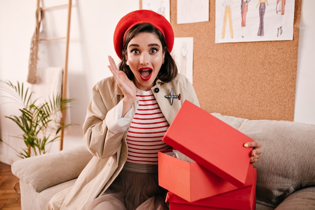 Überraschte Frau öffnet Geschenkbox und schaut in die Kamera. Schockiertes wundervolles Mädchen in moderner Herbstkleidung freut sich über ihr Geschenk.