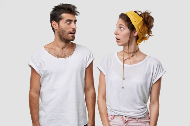 Überraschte Frauen und Männer diskutieren etwas Unglaubliches, schauen mit weit geöffneten Augen, haben einen überraschenden Gesichtsausdruck, tragen ein lässiges weißes T-Shirt, Model Indoor