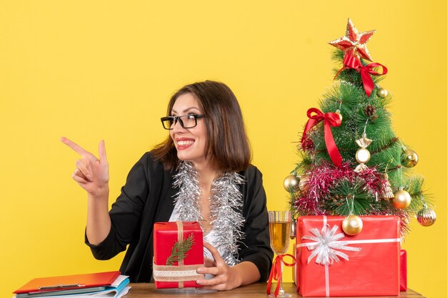 Überraschte Geschäftsdame im Anzug mit Brille, die ihr Geschenk zeigt, das etwas auf die rechte Seite zeigt und an einem Tisch mit einem Weihnachtsbaum darauf im Büro sitzt