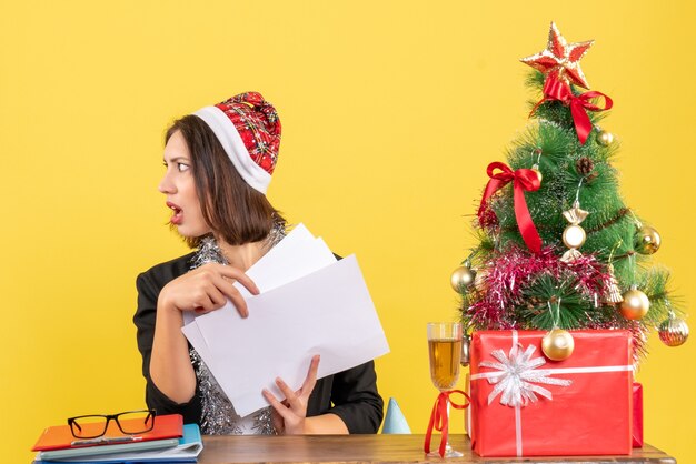 Überraschte Geschäftsdame im Anzug mit Weihnachtsmannhut und Neujahrsdekorationen, die Dokumente halten, die etwas betrachten und an einem Tisch mit einem Weihnachtsbaum darauf im Büro sitzen