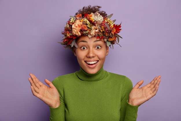 Überraschte glückliche ahnungslose Frau breitet Palmen aus, lächelt freudig, zeigt weiße Zähne, trägt handgemachten Herbstkranz und grünen Rollkragenpullover, isoliert über lila Hintergrund