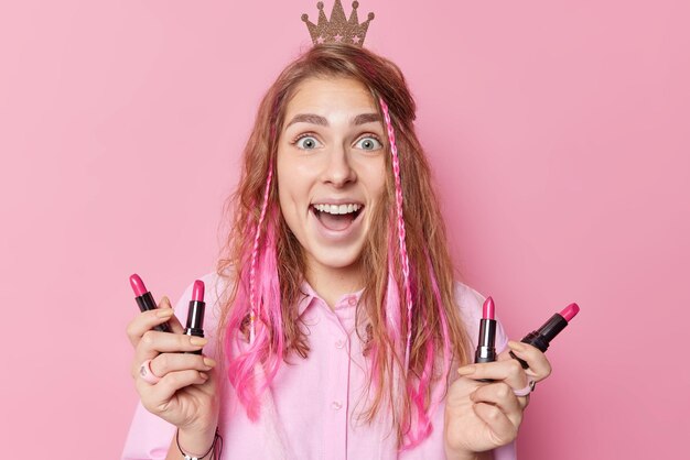 Überraschte glückliche Europäerin mit langen Haaren hält Lippenstifte wählt bestes Produkt bereitet sich auf Party vor trägt kleine Krone hat erstaunten Ausdruck dabei, sich isoliert über rosa Hintergrund zu schminken