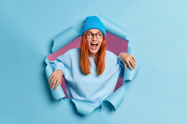 Überraschte glückliche Frau sieht mit großem Interesse aus, hält den Mund offen, trägt einen blauen Hut und ein Sweatshirt bricht durch das Papierloch