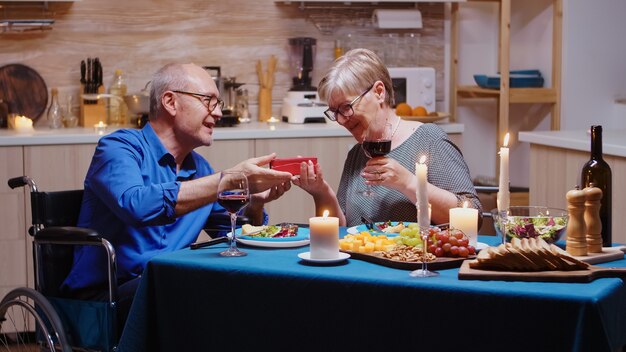 Überraschte Großmutter, die das Geschenk des Großvaters beim festlichen Abendessen betrachtet. Imobilisierter, gelähmter, behinderter älterer Ehemann, der mit seiner Frau zu Hause isst, das Essen genießt, ihren Jahrestag feiert