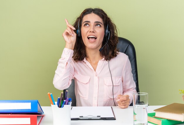 Überraschte hübsche kaukasische Call-Center-Betreiberin auf Kopfhörern, die am Schreibtisch sitzen und Bürowerkzeuge einzeln auf grüner Wand suchen und zeigen?