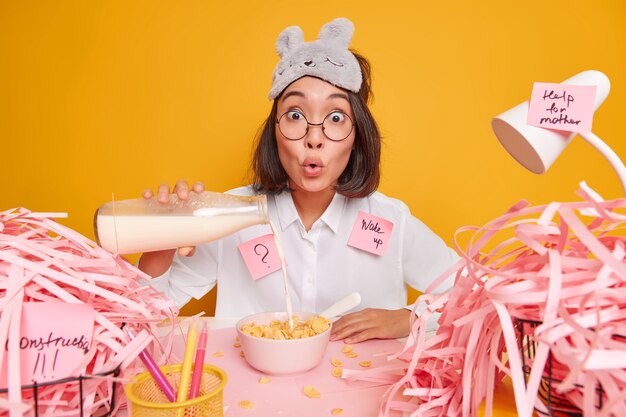 Überraschte junge Asiatin gießt Milch in Cornflakes, bereitet das Frühstück vor, macht Memo-Aufkleber durcheinander auf dem Desktop isoliert über gelber Wand hat einen vollen Terminkalender