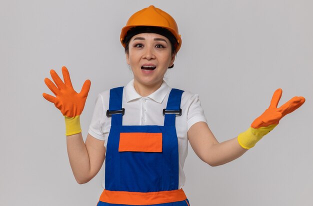 Überraschte junge asiatische Baumeisterin mit orangefarbenem Schutzhelm und Sicherheitshandschuhen, die die Hände offen halten