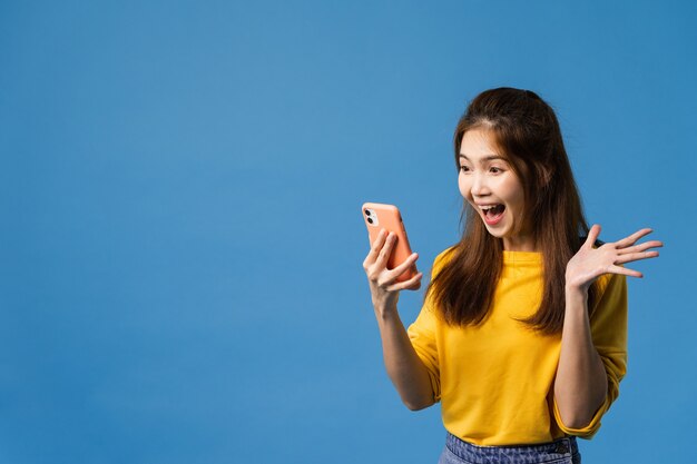 Überraschte junge asiatische Dame, die Handy mit positivem Ausdruck verwendet, lächelt breit, gekleidet in Freizeitkleidung und steht isoliert auf blauem Hintergrund. Glückliche entzückende frohe Frau freut sich über Erfolg.