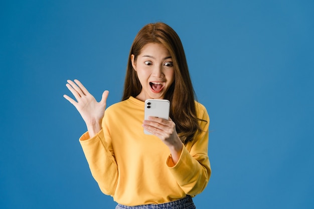Überraschte junge asiatische Dame, die Handy mit positivem Ausdruck verwendet, lächelt breit, gekleidet in Freizeitkleidung und steht isoliert auf blauem Hintergrund. Glückliche entzückende frohe Frau freut sich über Erfolg.