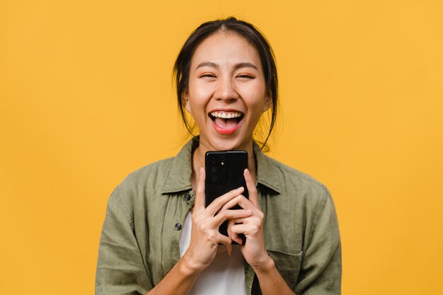 Überraschte junge asiatische Dame mit Handy mit positivem Ausdruck, breites Lächeln, gekleidet in Freizeitkleidung an gelber Wand. Glückliche entzückende frohe Frau freut sich über Erfolg.