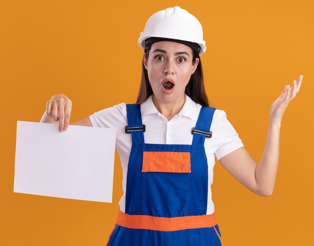 Überraschte junge Baumeisterfrau in Uniform, die Papierverbreitungshand lokalisiert auf orange Wand hält