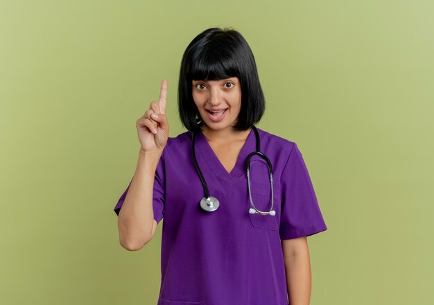 Überraschte junge brünette Ärztin in Uniform mit Stethoskop zeigt nach oben