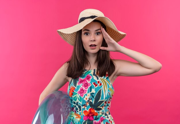 Überraschte junge Frau, die Hut hält, der Schwimmring hält und Gesicht mit Hand auf isolierter rosa Wand berührt