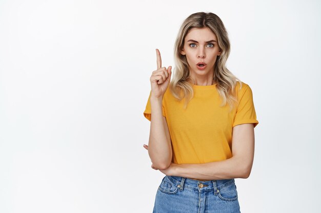 Überraschte junge Frau, die mit dem Finger nach oben zeigt und beeindruckt in die Kamera schaut, die gelbes T-Shirt und weißen Jeanshintergrund trägt