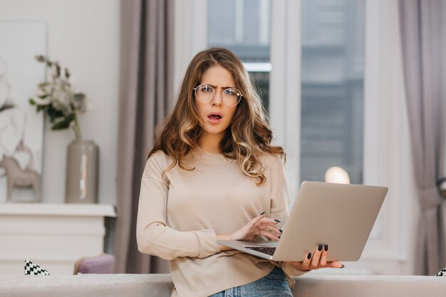 Überraschte junge Frau in beige Kleidung, die Laptop hält