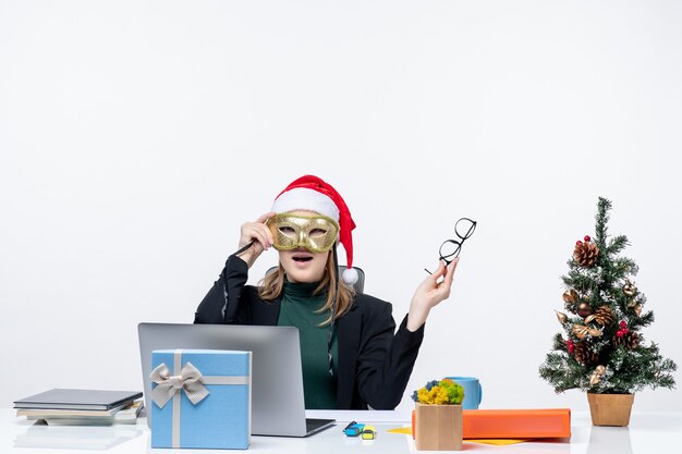 Überraschte junge Frau mit Weihnachtsmannhut, der Brillen hält und Maske trägt, die an einem Tisch mit einem Weihnachtsbaum und einem Geschenk darauf im Büro auf weißem Hintergrund sitzt