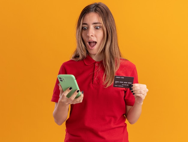 Überraschte junge hübsche Frau hält Kreditkarte und betrachtet Telefon isoliert auf orange Wand