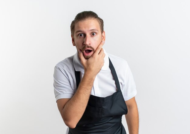 Überraschte junge hübsche Friseur, die Uniform trägt Hand auf sein Kinn isoliert auf weißer Wand trägt