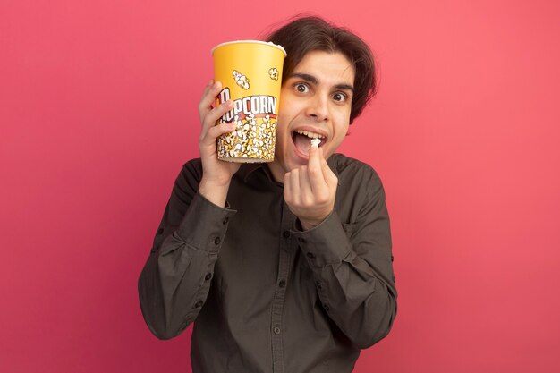 Überraschte junge hübsche Kerl, die schwarzes T-Shirt hält Eimer Popcorn mit Popcorn Frieden auf rosa Wand isoliert