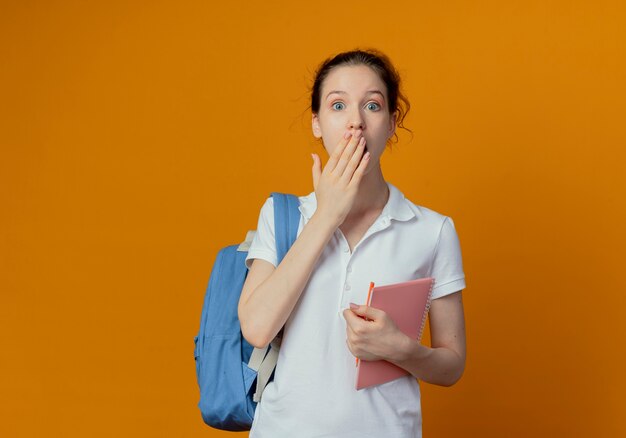 Überraschte junge hübsche Studentin, die Rückentasche hält, die Notizblock und Stift hält Hand auf Mund lokalisiert auf orange Hintergrund mit Kopienraum