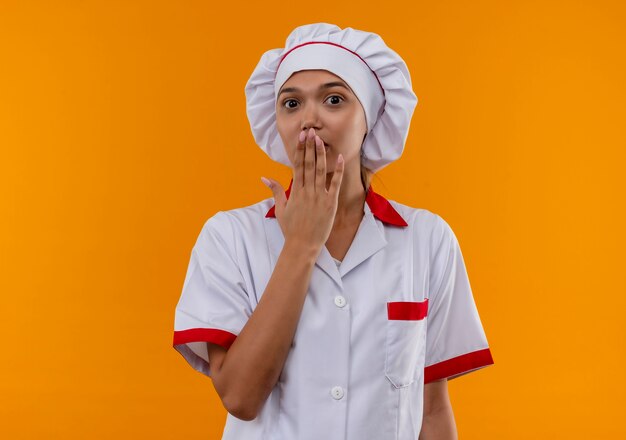 Überraschte junge Köchin, die Kochuniform trägt, bedeckte Mund mit Hand auf lokalisiertem orange Hintergrund mit Kopienraum