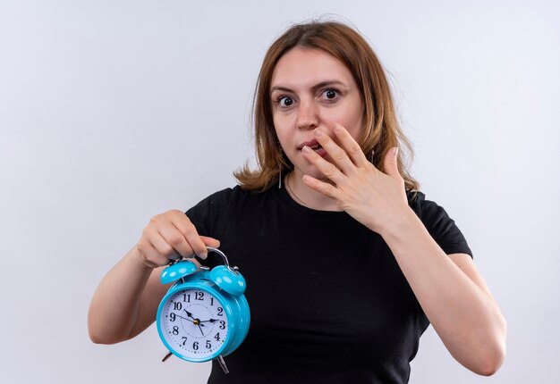 Überraschte junge lässige Frau, die Wecker hält und Hand auf Mund auf isolierte weiße Wand setzt