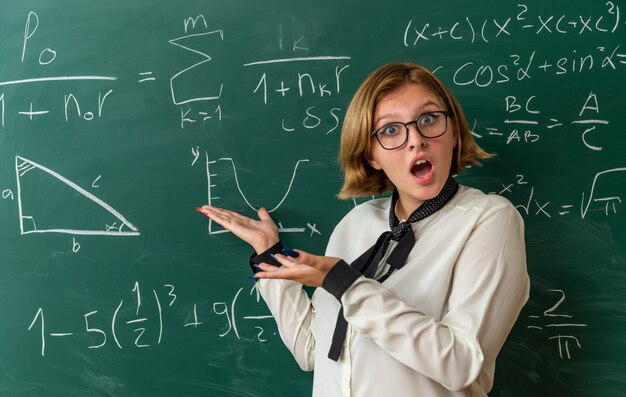 Überraschte junge Lehrerin mit Brille, die vor der Tafel steht, zeigt mit der Hand an der Tafel im Klassenzimmer