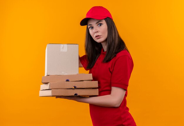 Überraschte junge Lieferfrau, die rotes T-Shirt in der roten Kappe hält, die eine Box mit Pizzaschachtel auf isolierter gelber Wand hält