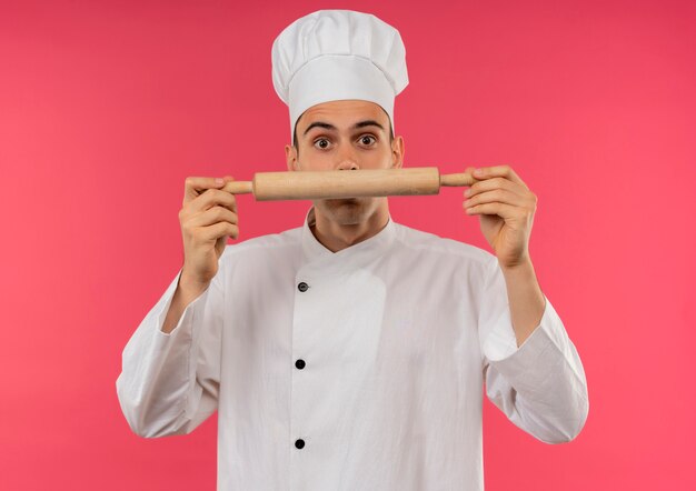 Überraschte junge männliche Köchin, die Kochuniform trägt, die Nudelholz um Gesicht hält