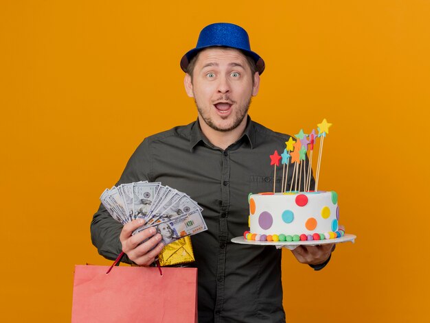 Überraschte junge Partei Kerl, der blauen Hut hält Kuchen mit Geschenktüten und Bargeld lokalisiert auf Orange trägt