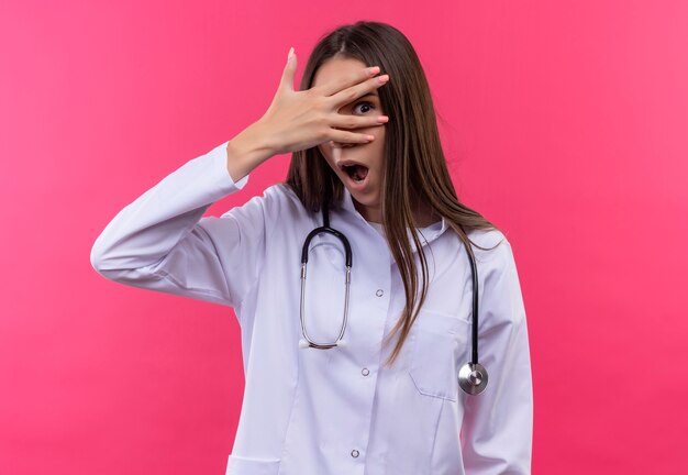 Überraschte junge Ärztin, die das medizinische Kleid des Stethoskops trug, bedeckte Auge mit Hand auf lokalisiertem rosa Hintergrund