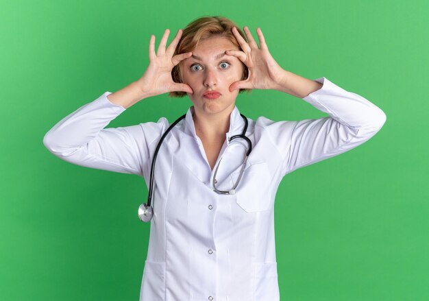 Überraschte junge Ärztin, die ein medizinisches Gewand mit offenen Augen des Stethoskops mit den Händen isoliert auf grünem Hintergrund trägt