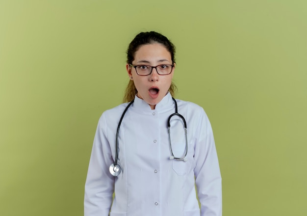 Überraschte junge Ärztin, die medizinische Robe und Stethoskop mit den auf olivgrüner Wand isolierten Gläsern trägt