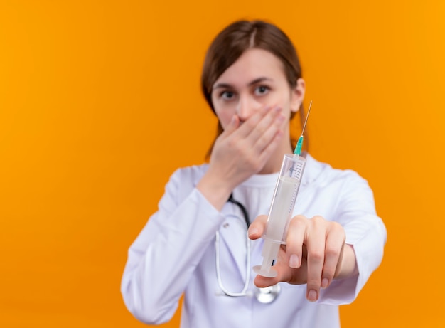 Überraschte junge Ärztin, die medizinische Robe und Stethoskop-Streckspritze trägt und Hand auf Mund auf isolierte orange Wand mit Kopienraum legt