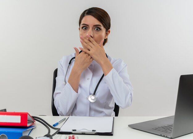 Überraschte junge Ärztin, die medizinische Robe und Stethoskop trägt, sitzt am Schreibtisch mit medizinischen Werkzeugen und Laptop, die Hände auf Mund lokalisiert auf weißer Wand setzen