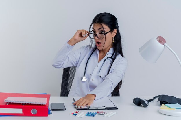 Überraschte junge Ärztin, die medizinische Robe und Stethoskop und Brille trägt, die am Schreibtisch mit medizinischen Werkzeugen sitzt, die Hand auf Schreibtisch setzen, die Brille ergreift und Seite isoliert betrachtet