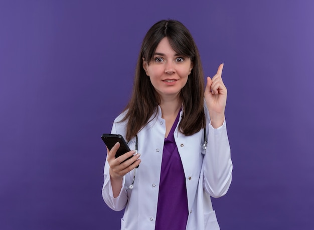 Überraschte junge Ärztin im medizinischen Gewand mit Stethoskop hält Telefon und zeigt auf isolierten violetten Hintergrund mit Kopienraum