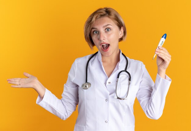 Überraschte junge Ärztin in medizinischem Gewand mit Stethoskop mit Thermometer isoliert auf orangem Hintergrund