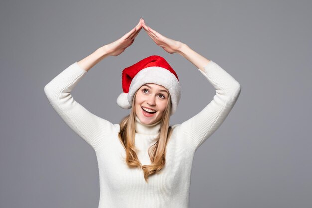 Überraschte junge Santa-Frau in gelbem Pullover, Weihnachtsmütze, die Hände über dem Kopf hält, wie das Dach des Hauses, das auf grauer Wand isoliert ist?