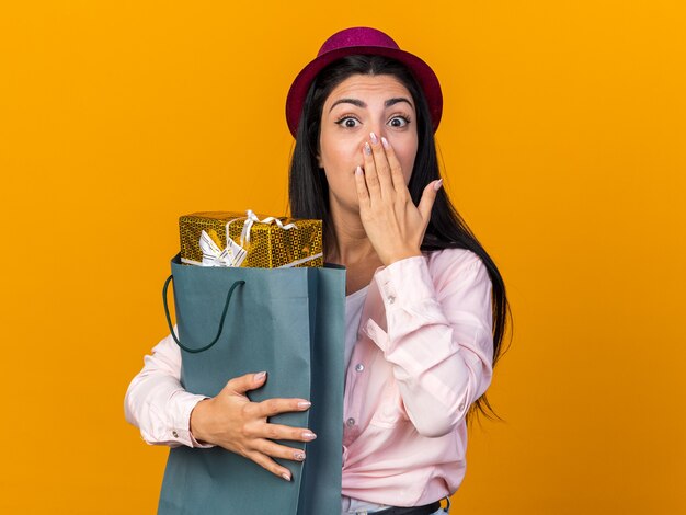 Überraschte junge schöne Frau mit Partyhut, die eine Geschenktüte hält, bedeckte den Mund mit der Hand isoliert auf der orangefarbenen Wand