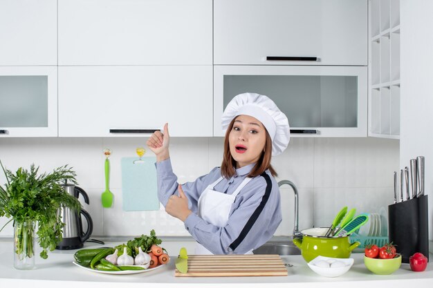 Überraschte Köchin und frisches Gemüse mit Kochutensilien und Okay-Geste auf der rechten Seite in der weißen Küche