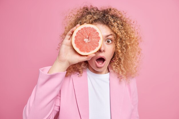 Überraschte, lockige europäische Frau bedeckt das Auge mit Grapefruitscheibe und hält den Mund offen, gekleidet in stilvolle Kleidung, isoliert über rosafarbener Wand. Sommer Erfrischung. Zitrusfrüchte Konzept.