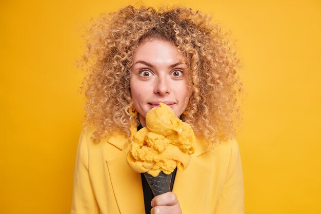 Überraschte lockige Frau isst leckeres Eis, kümmert sich nicht um Kalorien sieht mit beeindrucktem glücklichem Ausdruck einzeln über gelber Wand aus. Frau hat Eis zum Nachtisch