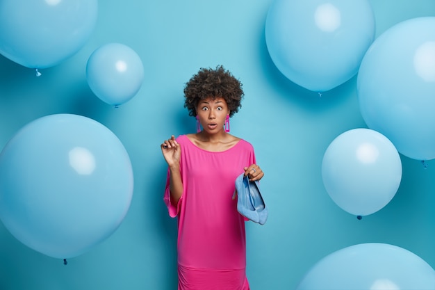 Überraschte lockige Frau wählt Outfit für perfekten Geburtstag, trägt in rosa festlichem Kleid und hält blaue Schuhe mit hohen Absätzen, merkt, dass sie vergessen hat, Tasche zu kaufen. Mode- und Feierkonzept.