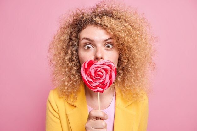 Überraschte lockige junge Frau bedeckt den Mund mit herzförmigen Süßigkeiten hat süße Zähne hält Lollipop gekleidet in gelber Jacke isoliert über rosa Wand. Viel Zucker ist nicht ok. Zuckerhaltige Leckereien